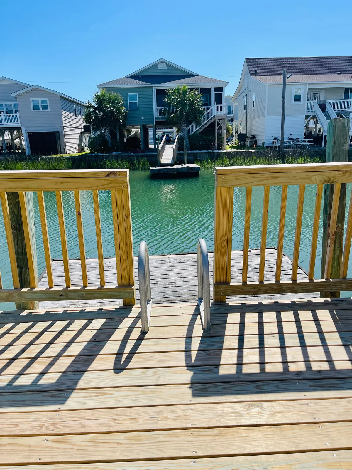 A serene wooden dock extending over calm green water, with palm trees and surrounding houses in view. The dock is a focal point for leisure activities like fishing, boating, or simply enjoying the tranquil atmosphere. The water reflects the sunset, enhancing the relaxing mood.