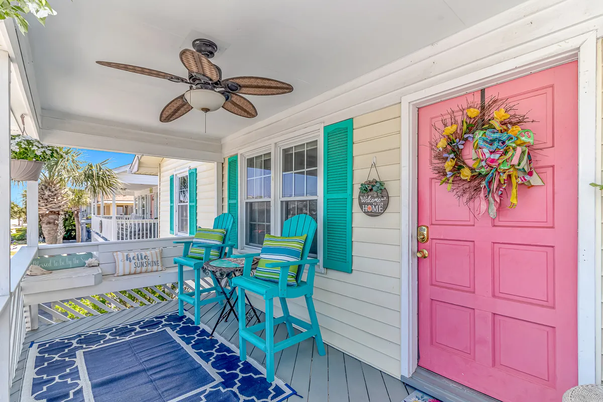 This exterior image features a welcoming front porch with a cheerful pink door, blue chairs, a decorative wreath, and a textured rug. The porch setting is perfect for enjoying morning coffee, reading, or socializing with neighbors. The overall mood is bright and inviting, suggesting a leisurely and upscale lifestyle.