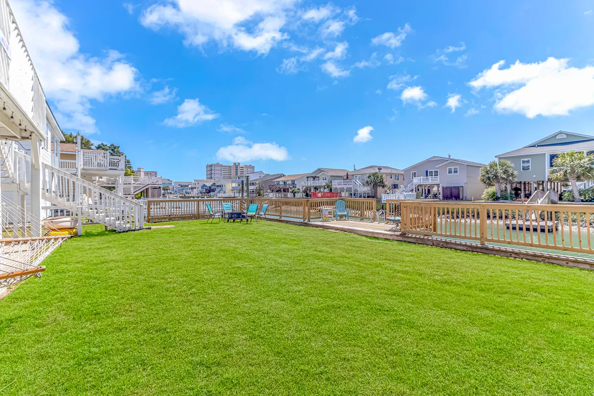 A vibrant backyard scene featuring a well-manicured green lawn bordered by a fenced deck area. Brightly colored lounge chairs are arranged for sunbathing or socializing, offering ample space for both relaxation and group activities. The area is open and inviting, perfect for pets to roam and families to gather.