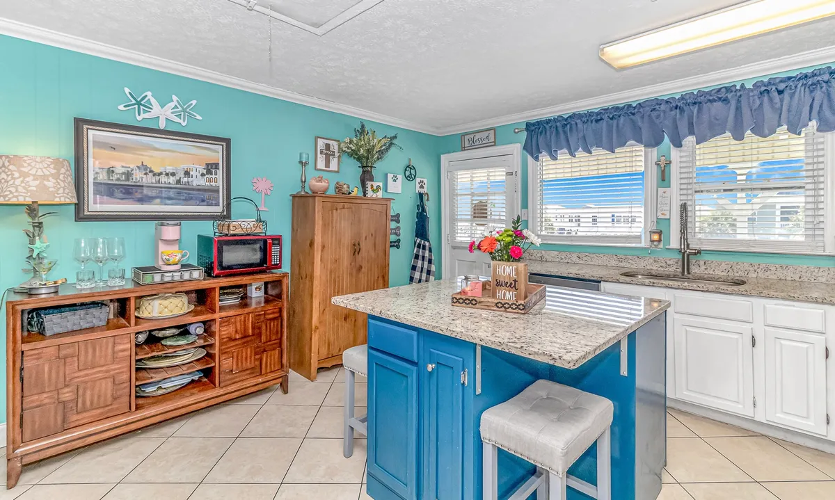 A cheerful kitchen scene with a granite countertop, blue island, decorative wall art, and a wooden cabinet. The space is vibrant and full of life, designed for both socializing and preparing meals, with ample counters and inviting seating.