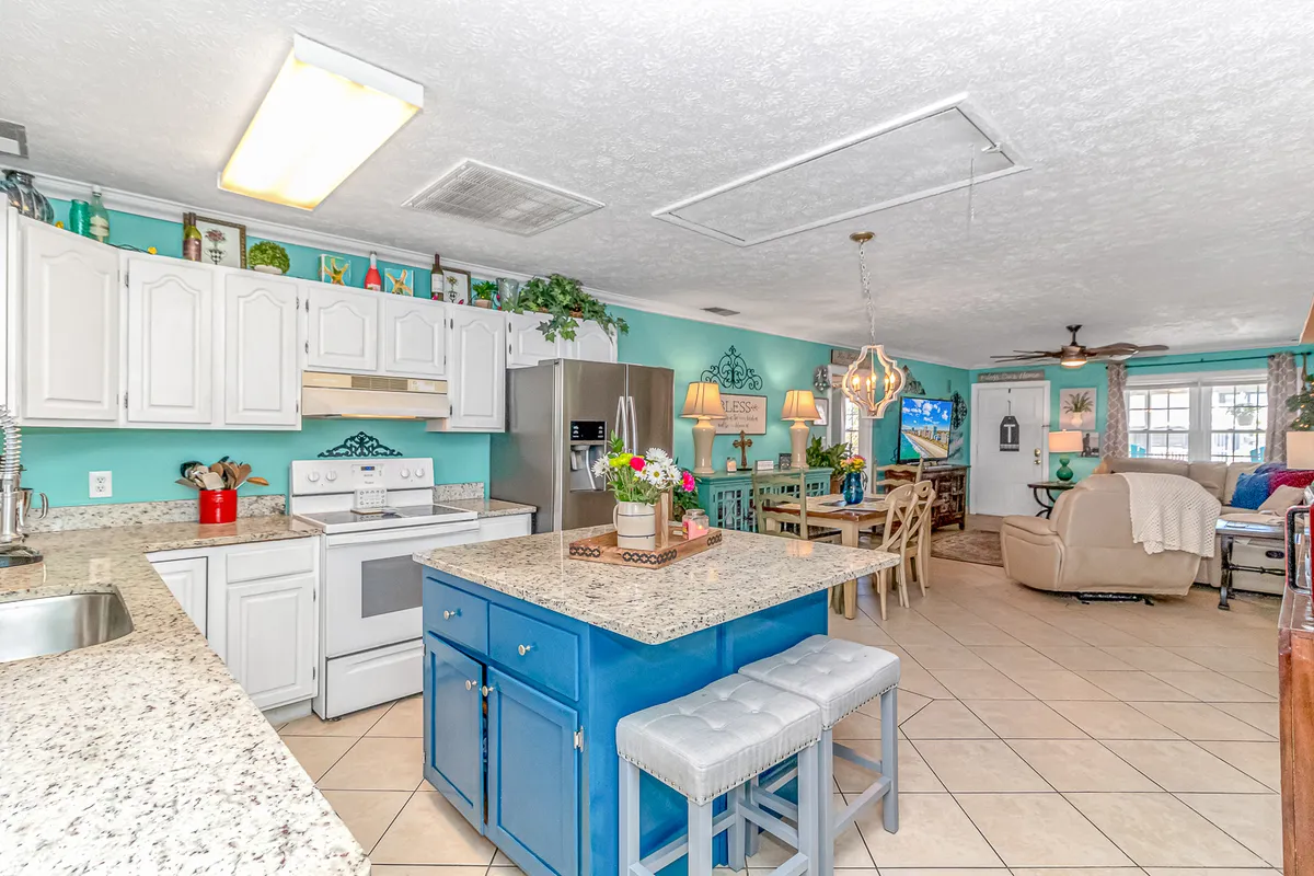 A modern dining area adjacent to a blue kitchen island, featuring granite countertops, white cabinetry, and decorative plants. The area is bathed in natural light, offering a welcoming space for meals and entertaining, with a layout that promotes easy flow between kitchen, dining, and living spaces.