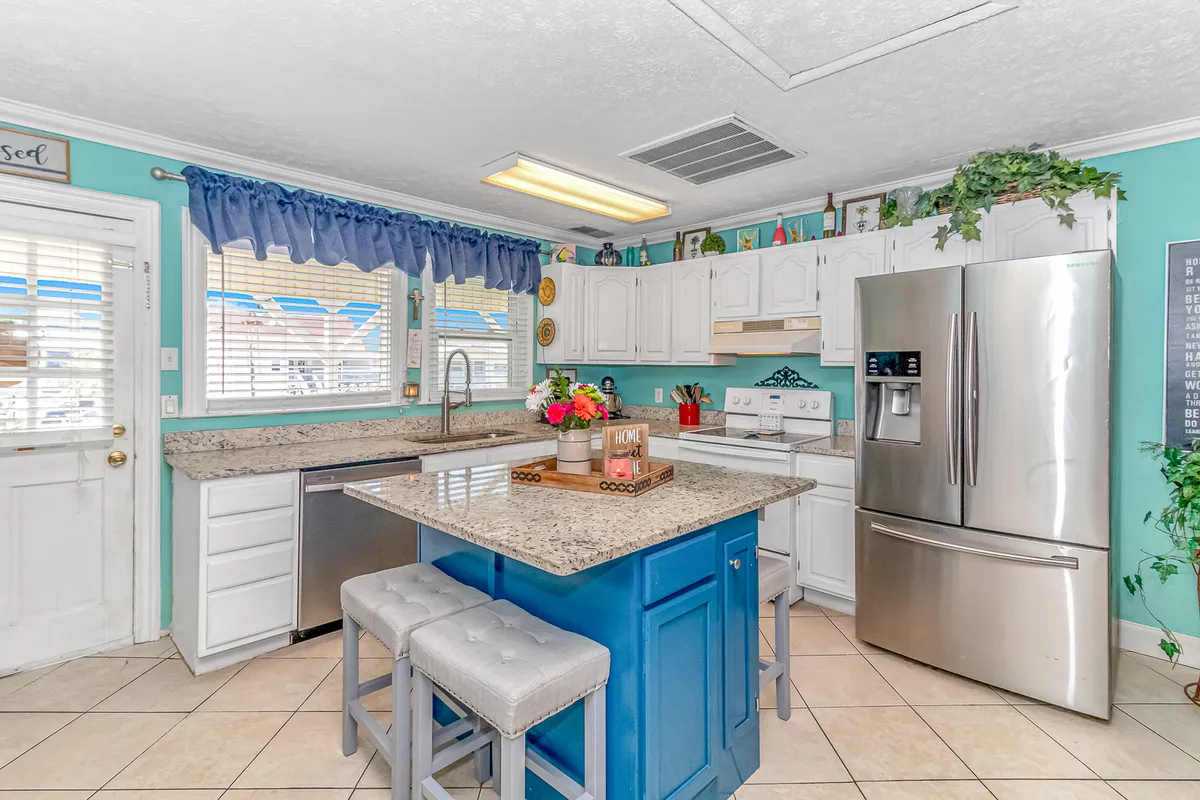 This image presents a warm and welcoming kitchen featuring a granite countertop, a blue kitchen island, crisp white cabinetry, stainless steel refrigerator, and decorative plants. The space is open and clearly designed for both cooking and gathering, creating an inviting heart-of-the-home atmosphere.