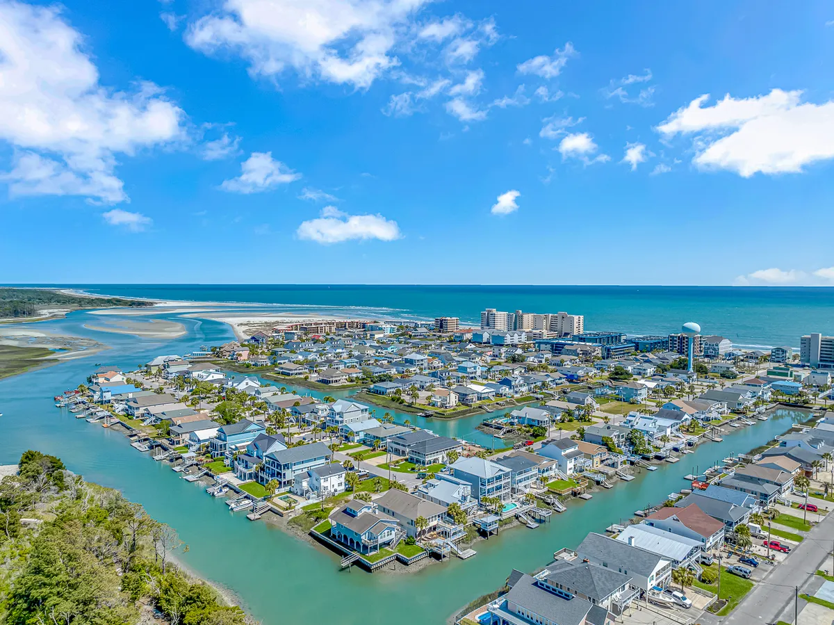 A panoramic view that captures coastal homes, canals, and a distant ocean horizon under a clear blue sky. The image emphasizes the area's pristine setting, easy access to both beach and water activities, and the overall laid-back coastal lifestyle.