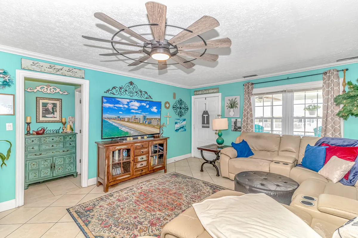 A cheerful living area with turquoise walls, wooden furniture, a large ceiling fan, vibrant artwork, and a textured rug. The space is filled with natural light, and the windows offer glimpses of the outdoors, enhancing the connection to the waterfront setting.