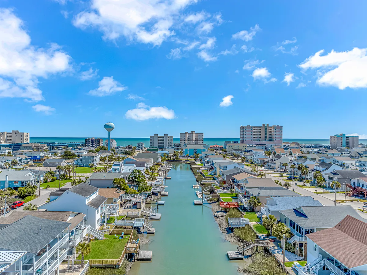 A scenic view of the canal lined with beachfront homes, a water tower in the distance, and boats moored along the waterway. The image conveys a sense of exploration, relaxation, and connection to both water and neighborhood life. Activities like boating, fishing, and evening walks are suggested by the setting.