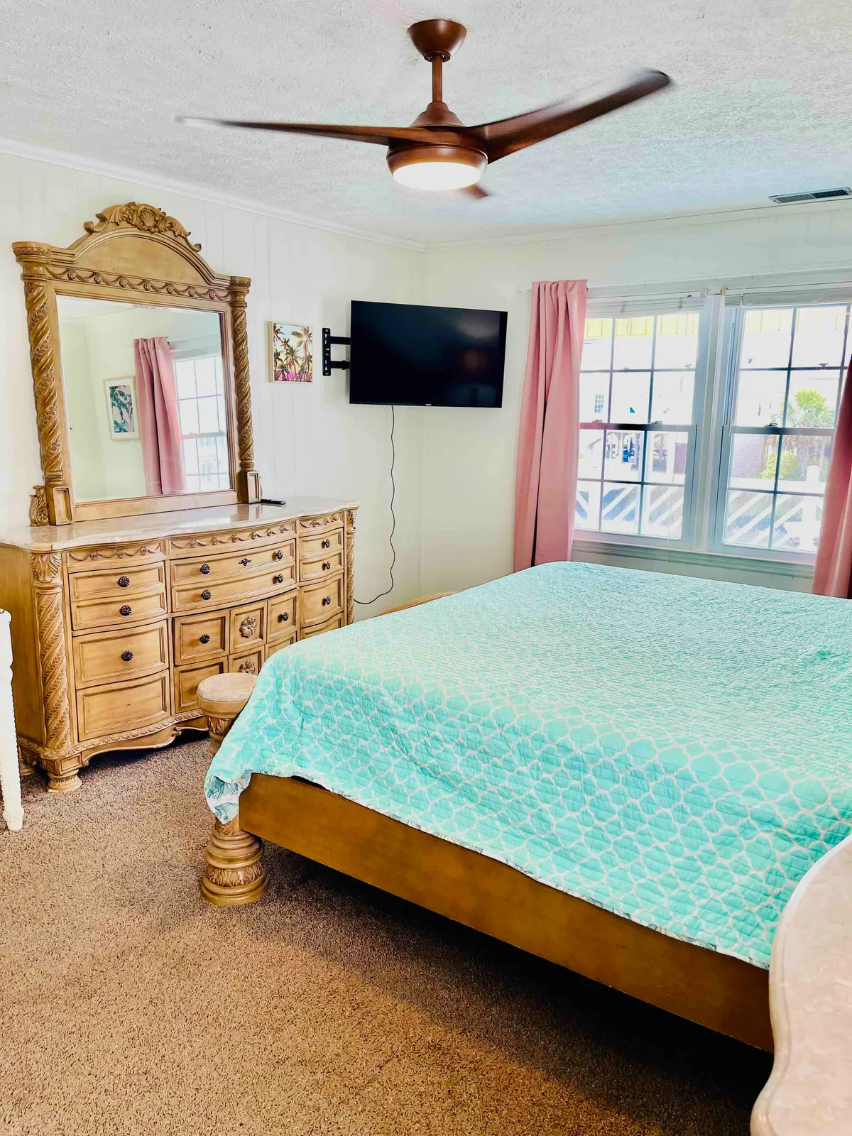 A spacious, comfortable bedroom with a bed dressed in a turquoise quilt, a large dresser and mirror, a wall-mounted TV, and pink curtains. The room exudes a cozy, serene vibe—perfect for peaceful mornings or unwinding at night.