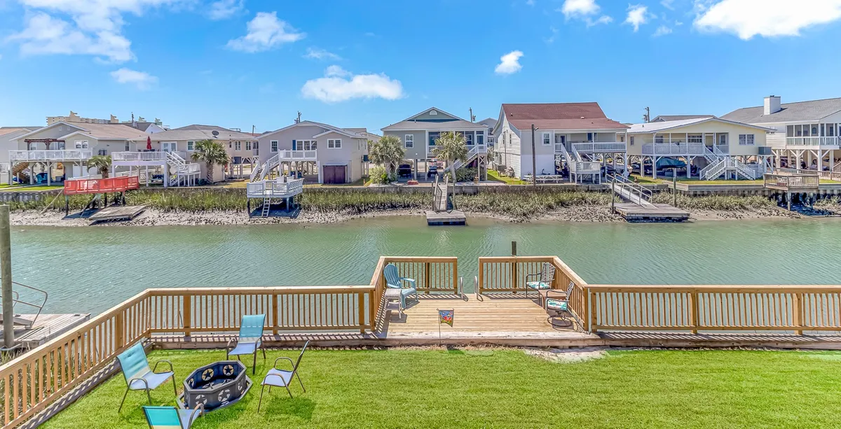 A waterfront deck with ample outdoor seating, a spacious lawn, and nearby palm trees. The setup is designed for family gatherings, sunbathing, fishing, or lounging by the water. Neighboring houses and a wide waterway create a picturesque backdrop.